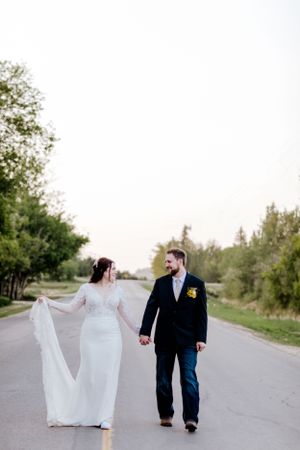 A couple walks hand in hand on a pathway with flowing wedding dress fabric.