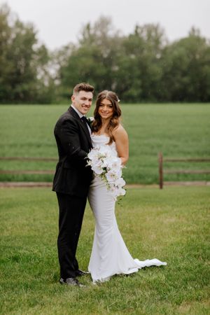 A newlywed couple poses with a white orchid bouquet in a green meadow.