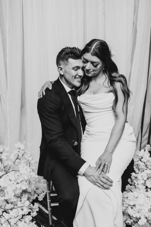 Black and white wedding photo with bride and groom embracing by floral arrangements.