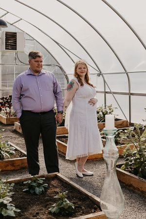 Newlyweds posing together in greenhouse surrounded by potted plants and garden beds.