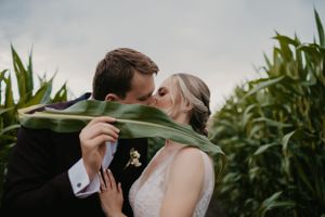 Newlyweds share a romantic kiss behind a large green leaf in a cornfield.