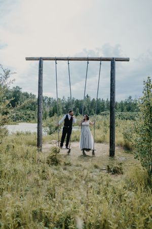 Bride and groom on rustic wooden swing set in a meadow by a lake.