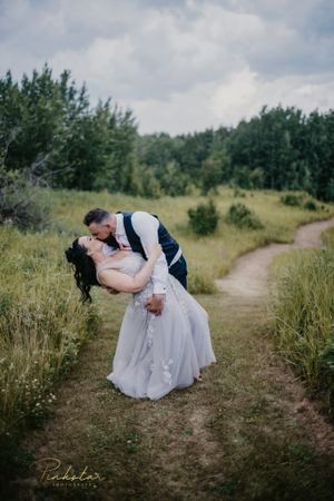 A romantic couple shares an intimate dip kiss on a winding dirt path surrounded by lush greenery.