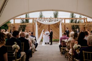 Wedding ceremony taking place under an elegant draped arch in a tent with guests seated on gilded chairs.