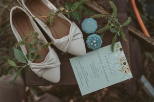 Wedding details flatlay with white bridal shoes, invitation and decorative elements on dark surface.