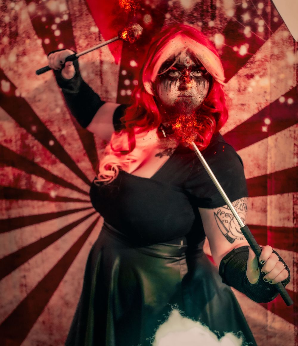 A dramatic costumed figure with red hair poses against a starburst background with props.