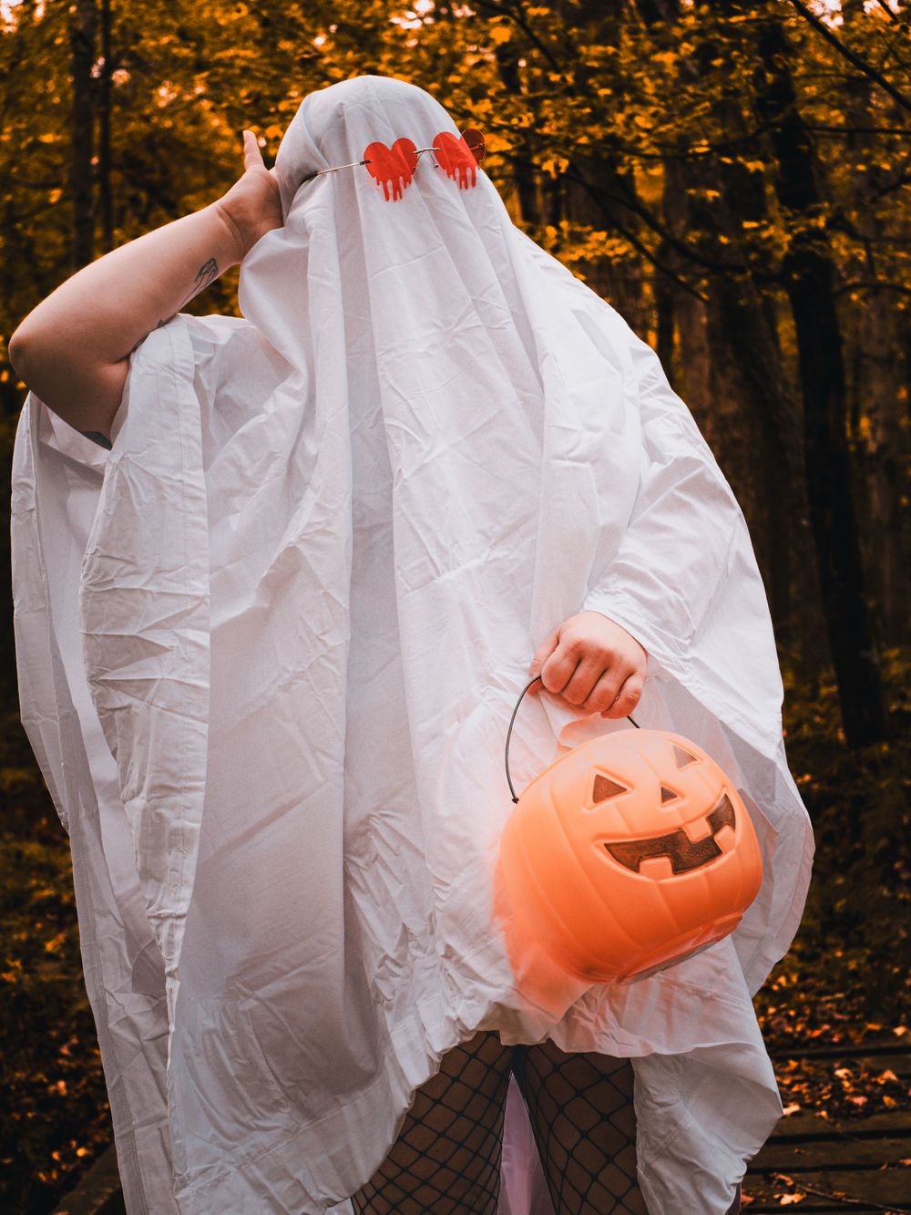 Ghost figure in white sheet holds orange Halloween pumpkin bucket against autumnal forest backdrop.
