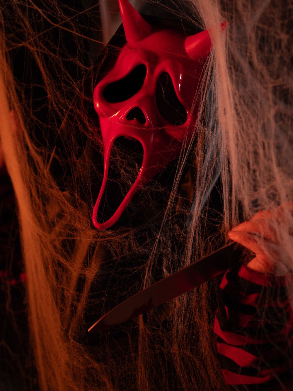 Red devil mask and knife among spider webs in dramatic orange lighting.