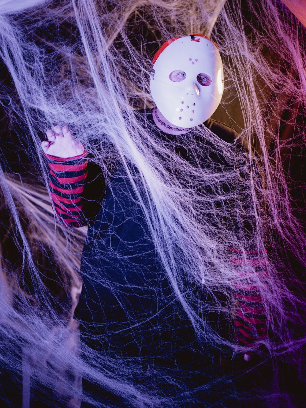 White hockey mask and striped sleeve emerging from spooky cobwebs with purple lighting.