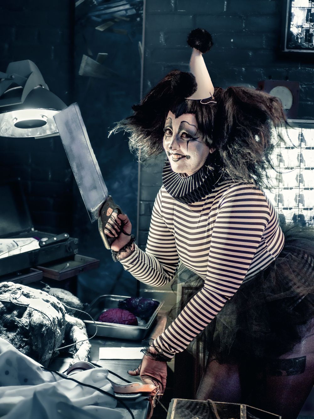 A performer in striped clothing and theatrical makeup examines props in a dark room.