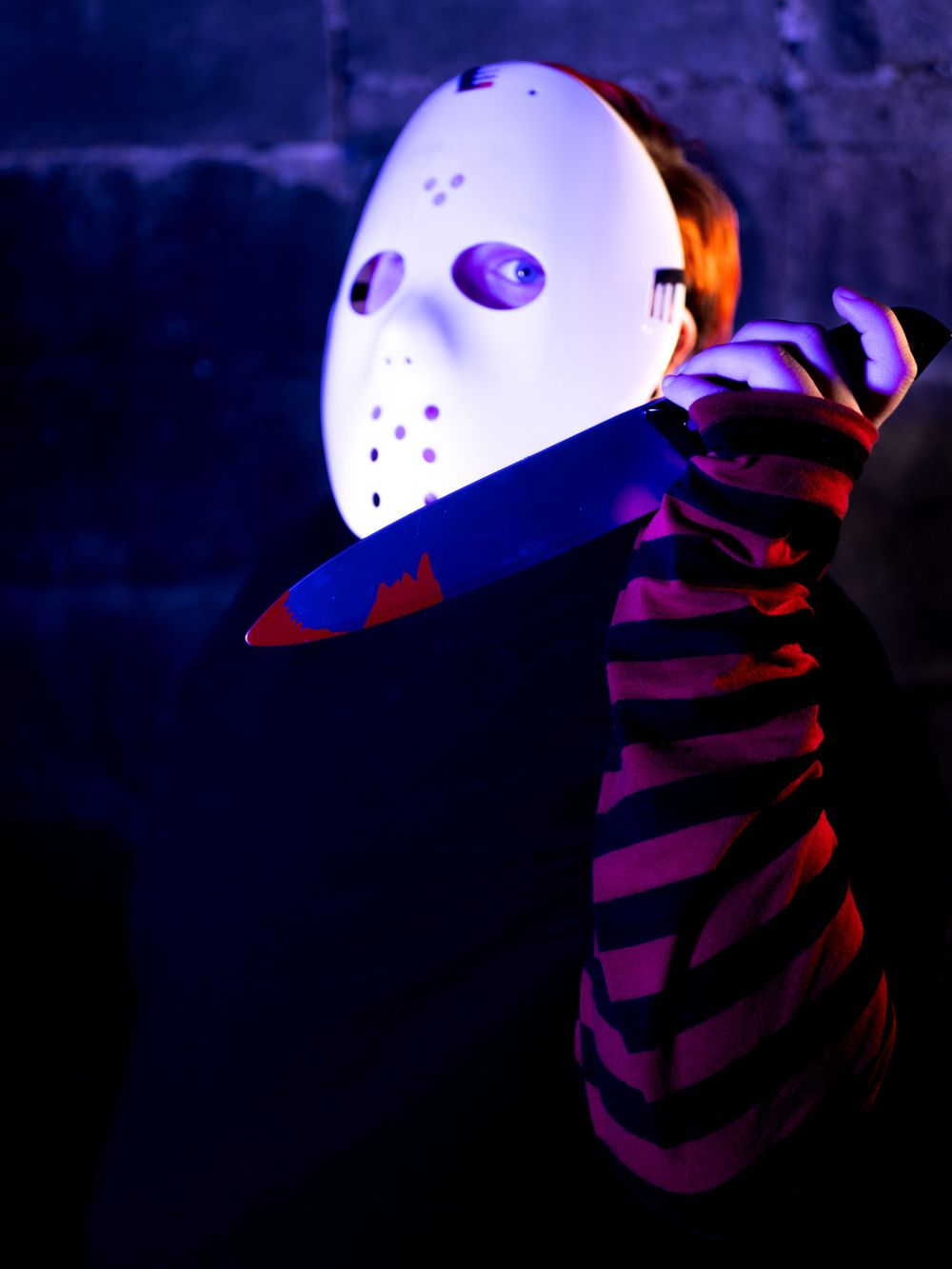 Hockey masked figure holding knife with striped sleeve in dark atmospheric lighting.