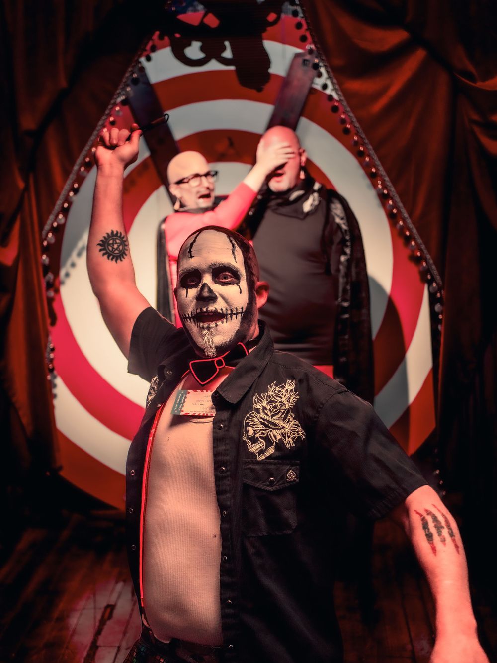 Performer with skull makeup in open vest against red and white circular backdrop.