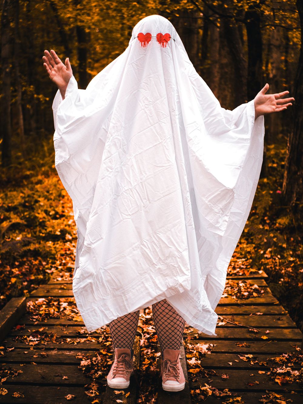 A ghost costume with red heart eyes poses with outstretched arms in an autumn forest setting.