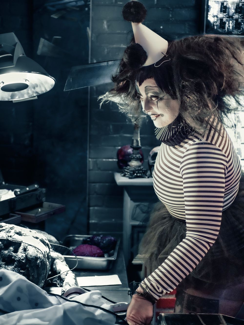 A theatrical character in striped clothing works at a prop table under dramatic lighting.