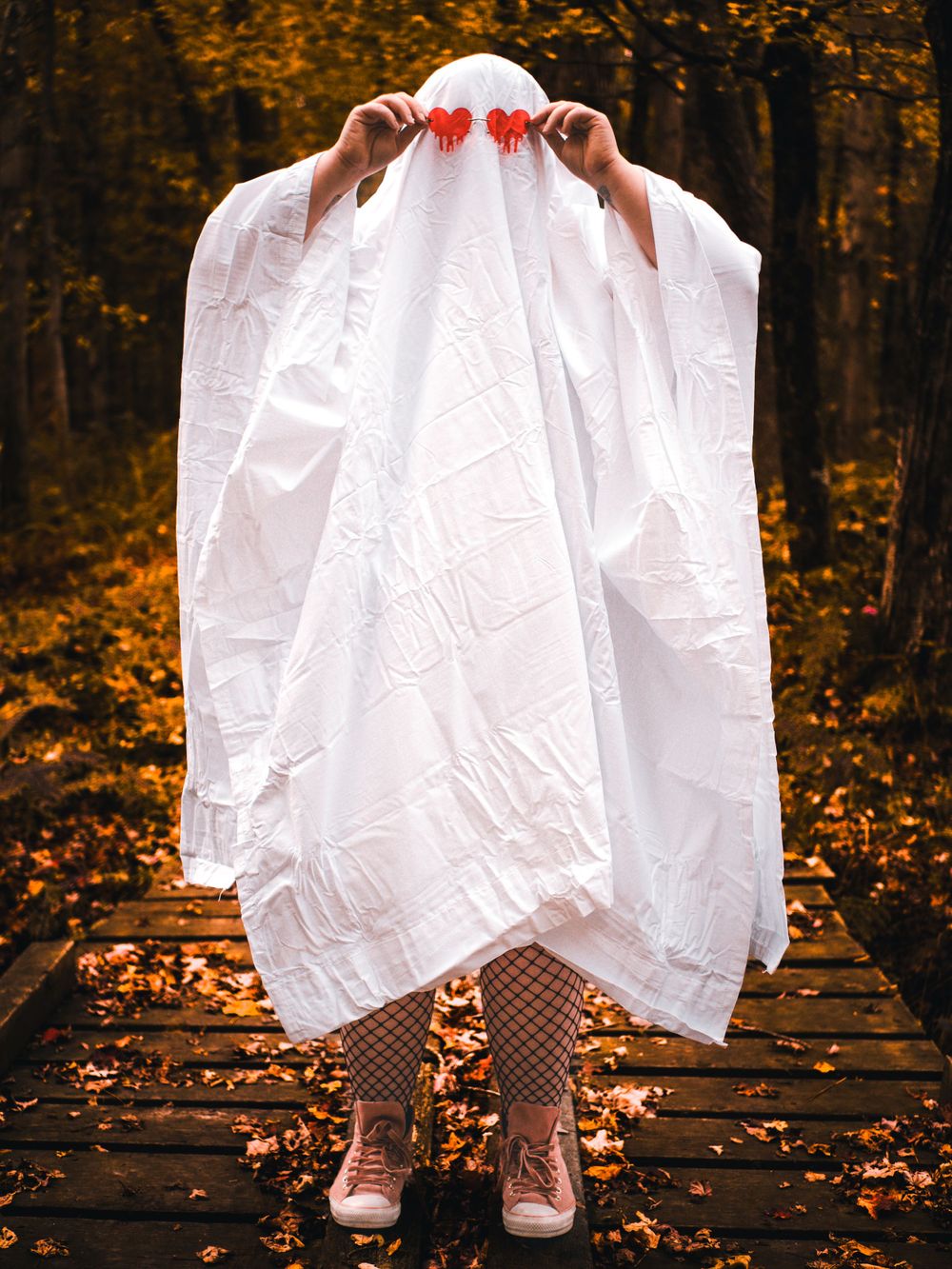 Ghost figure in white sheet and fishnet stockings holds their ghost eyes against fall foliage.