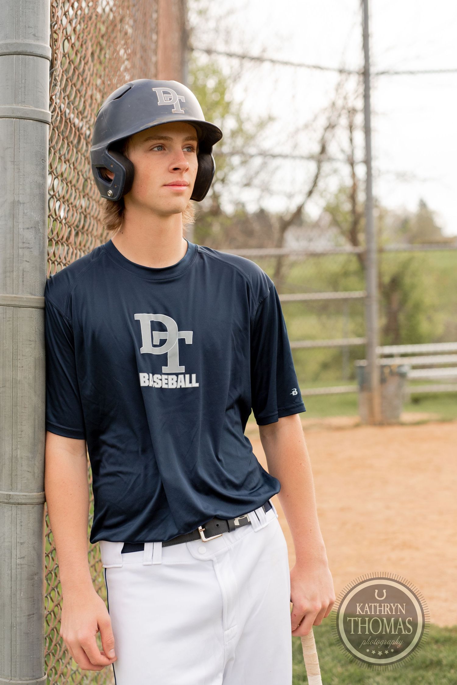 Baseball player in navy jersey and helmet stands against chain-link fence on baseball field.
