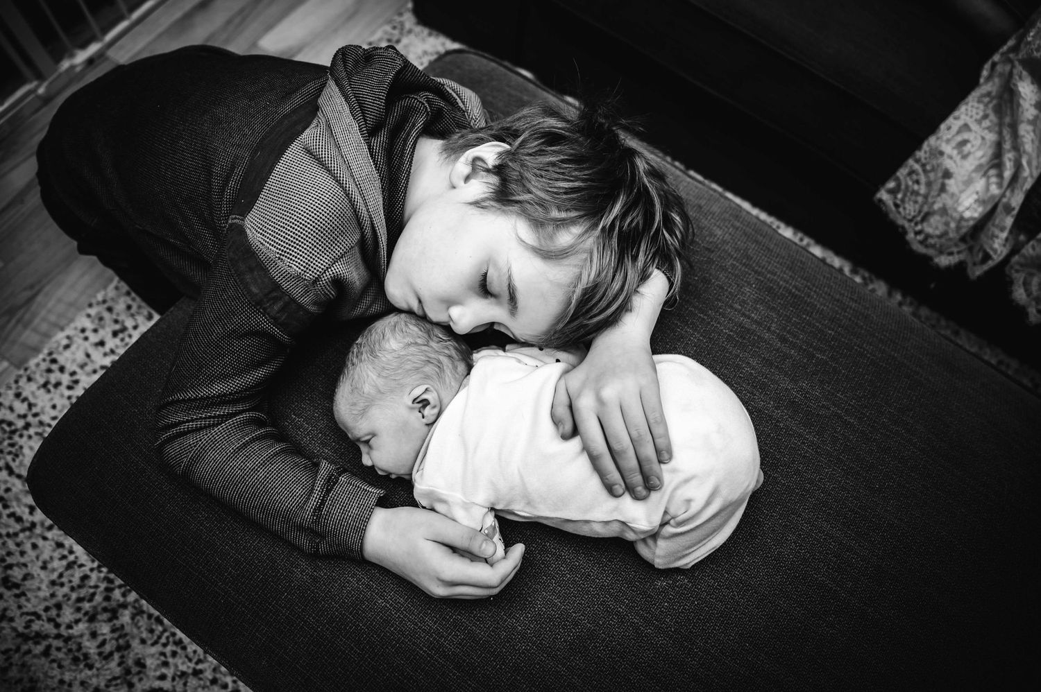 Black and white photo of a child tenderly cuddling a swaddled newborn baby on the floor.