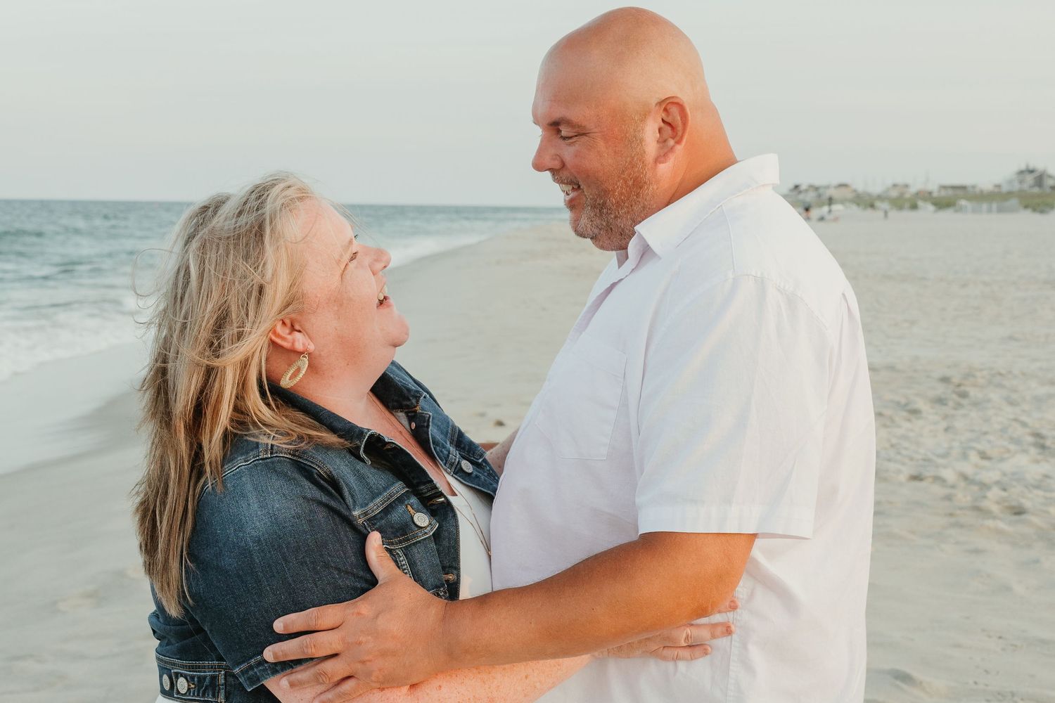 Romantic couple embracing on beach at sunset wearing white and denim.