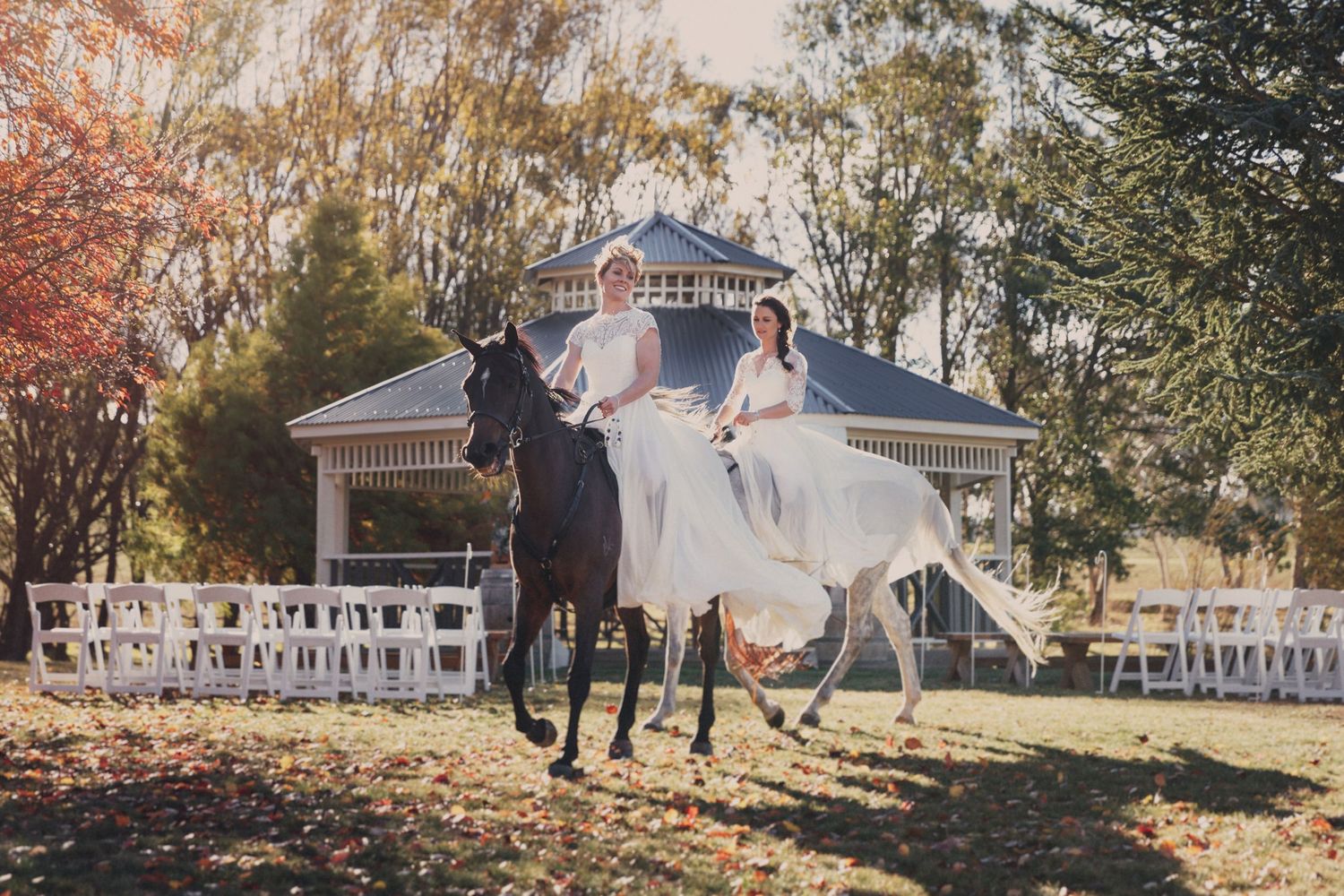 Bride and groom ride horses across autumn lawn with white gazebo and ceremony chairs in background.