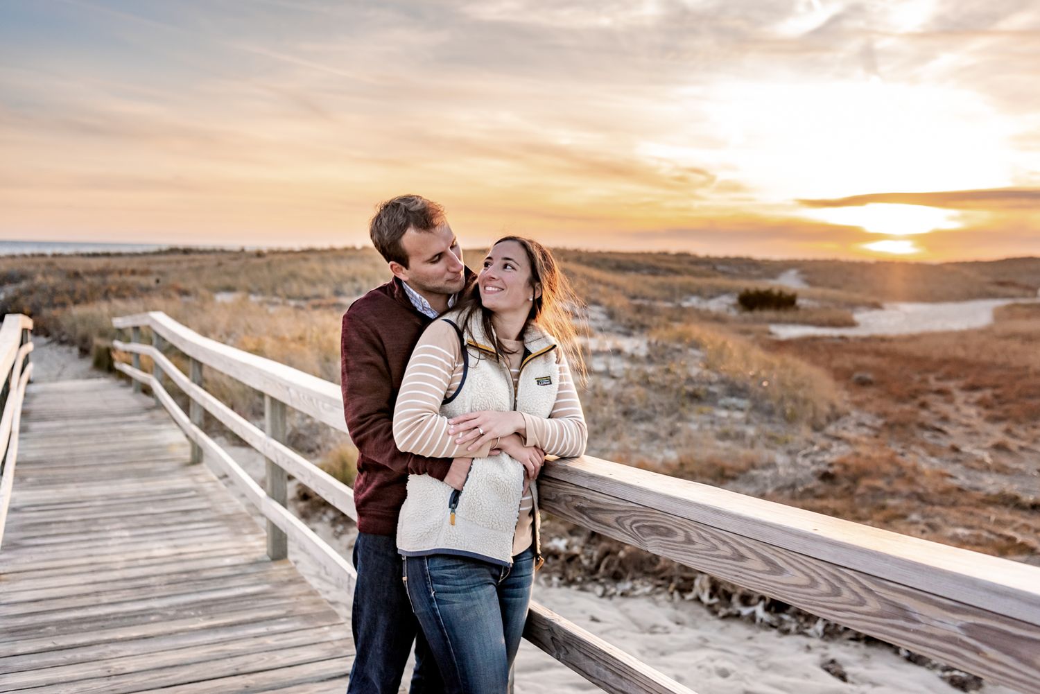 Romantic couple embracing on wooden boardwalk during golden sunset over coastal landscape in Rhode Island.