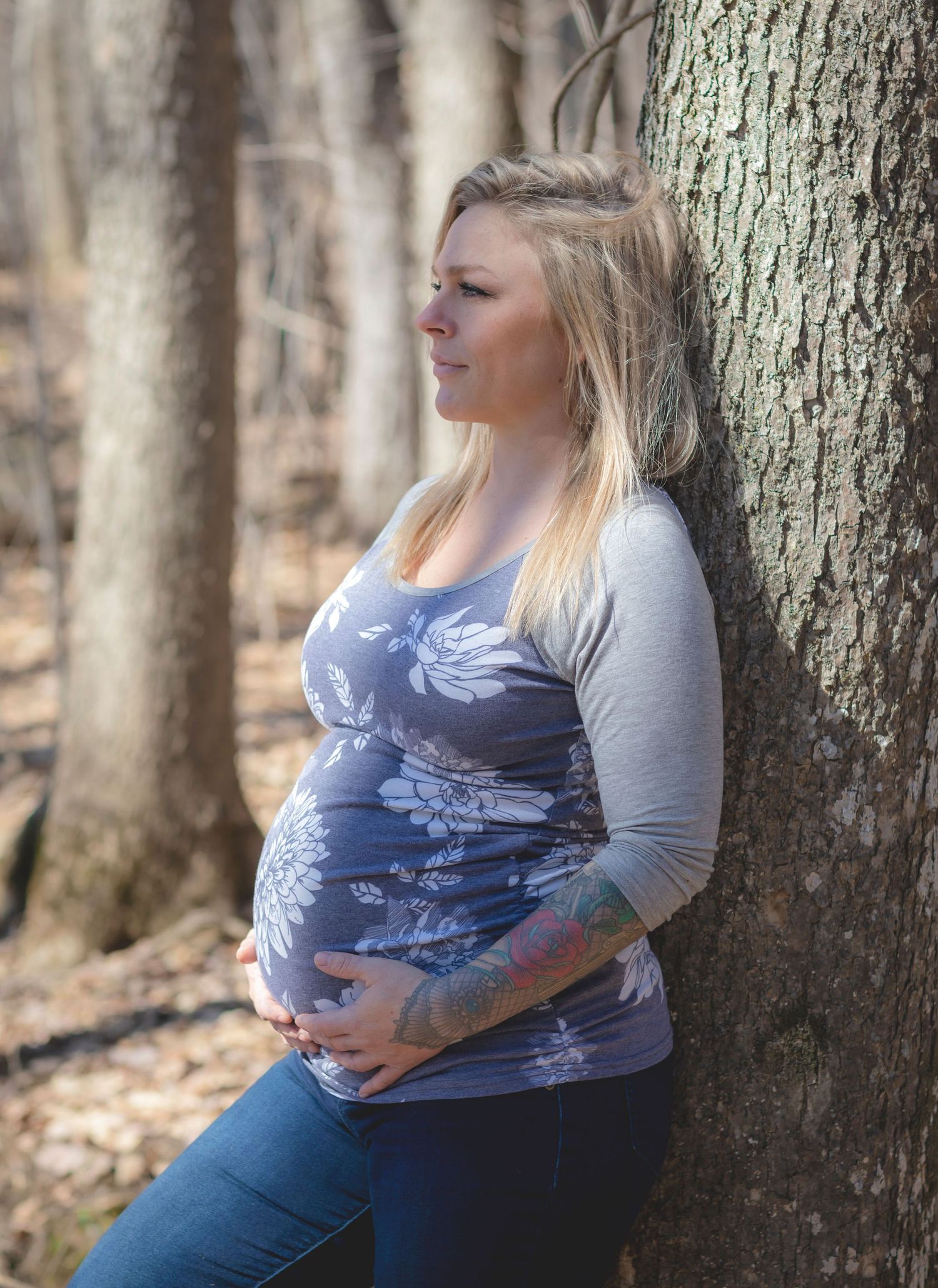 Maternity photo session showing an expectant mother in casual attire leaning against a tree trunk in a wooded setting.