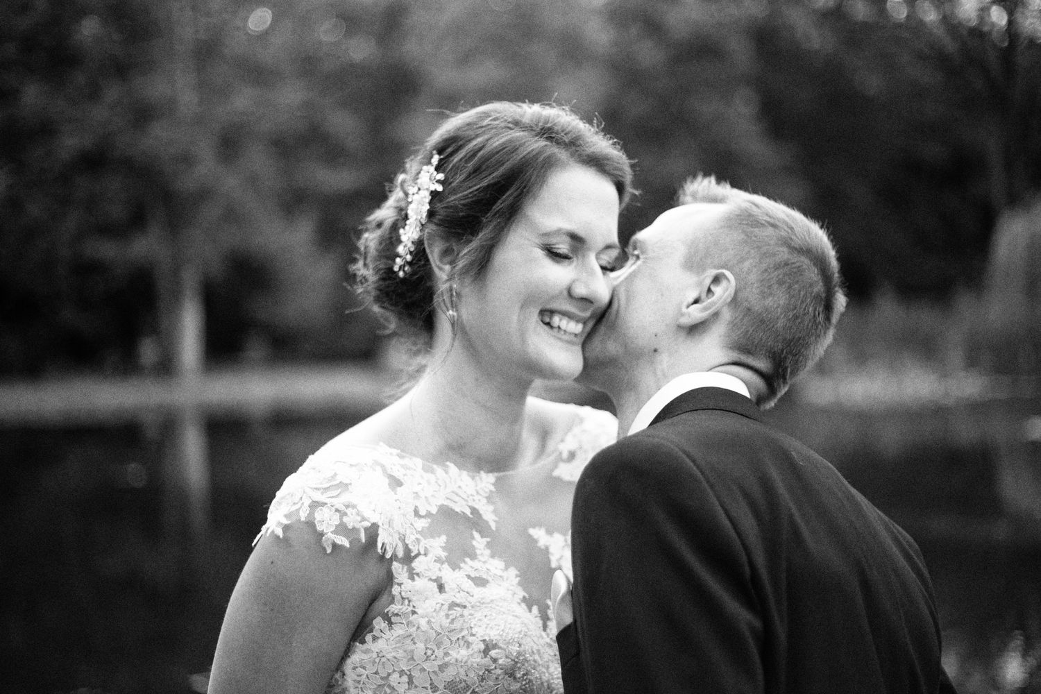 Black and white photo of a smiling bride being kissed on the cheek by a groom near a lake.