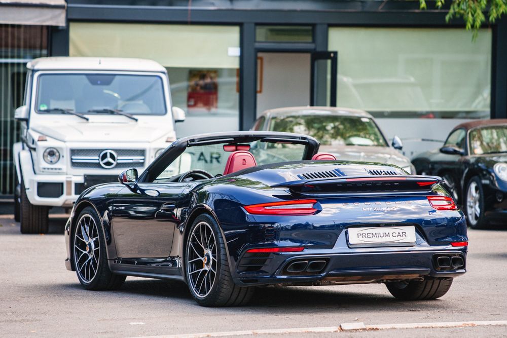 Rear three-quarter view of a navy Porsche 911 convertible with white Mercedes in background.
