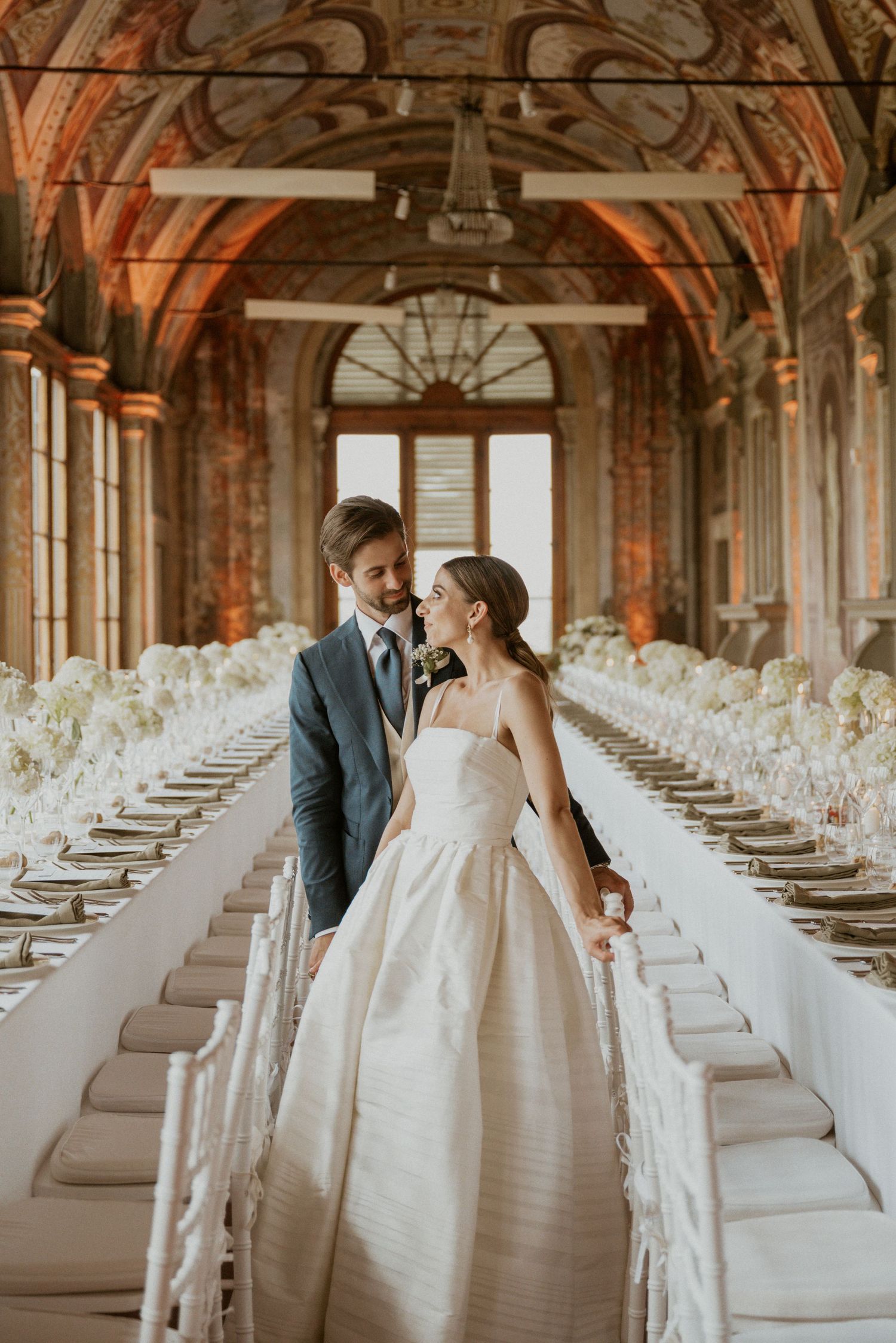 Wedding couple in an elegant hall with arched ceilings and rows of white chairs.