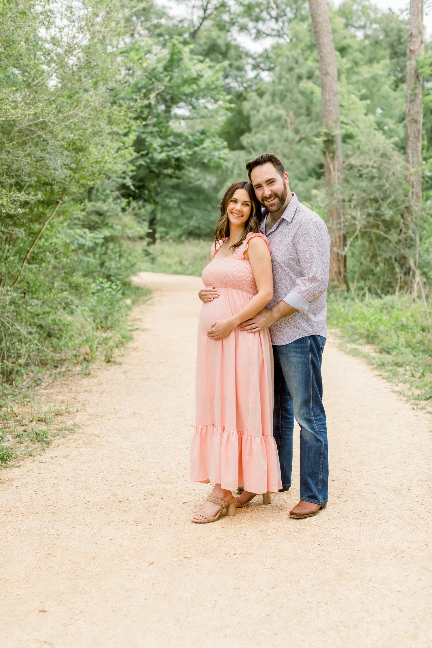 A romantic moment captured on a dirt path surrounded by trees with a couple embracing in casual attire.