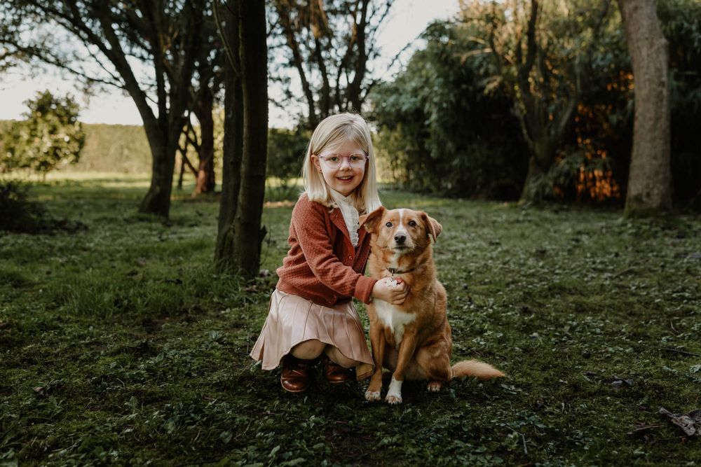 A child in a rust-colored jacket poses with a golden retriever dog in a wooded setting.