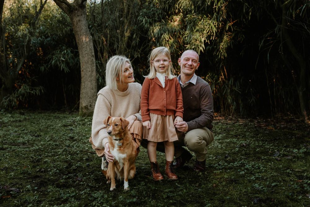A family photo in earthy tones with their golden retriever dog against a green garden backdrop.