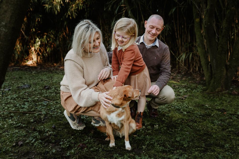 A family kneels together with their golden retriever pet in a lush garden setting.