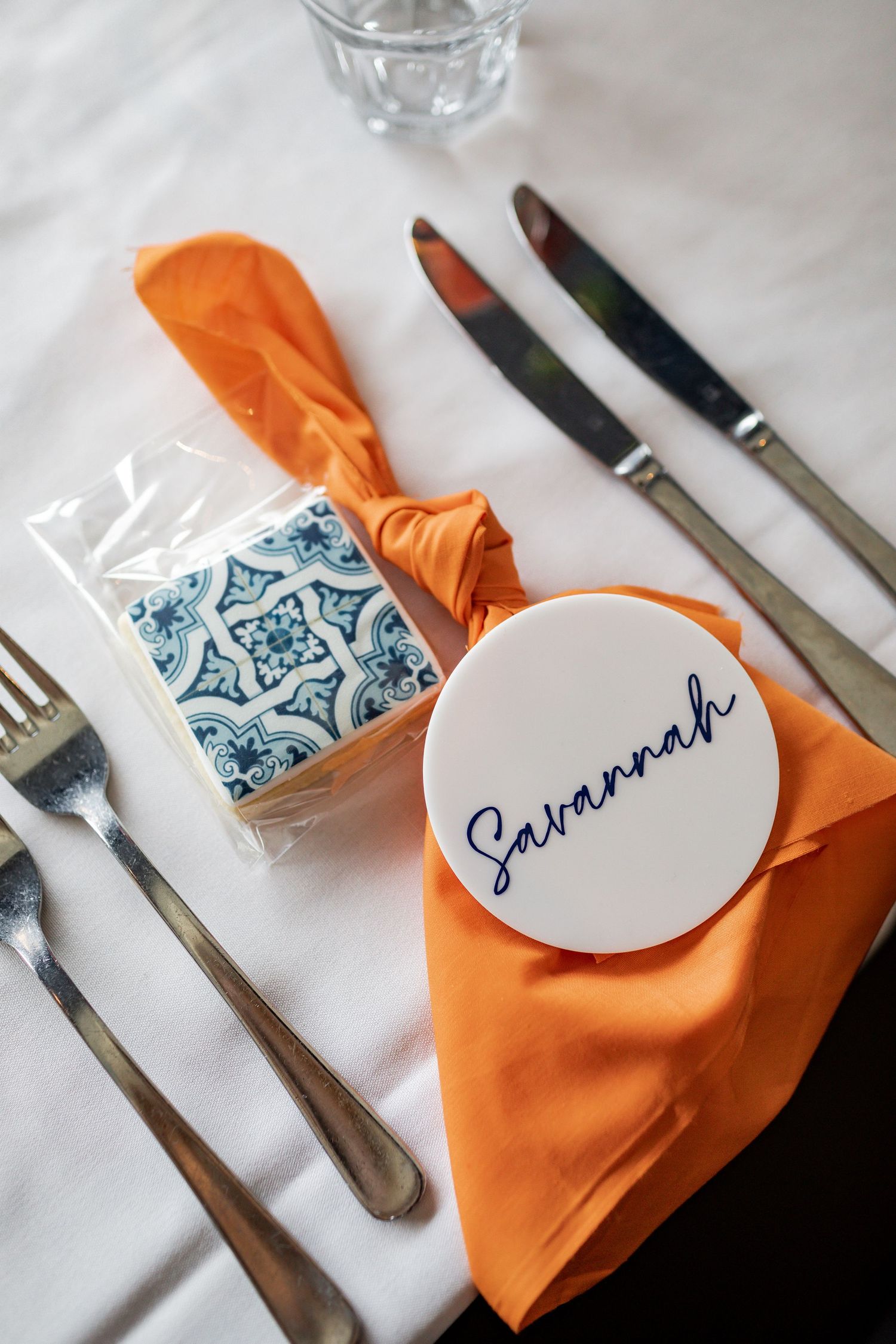 Place setting with orange napkin, patterned tile coaster, and personalized name tag.