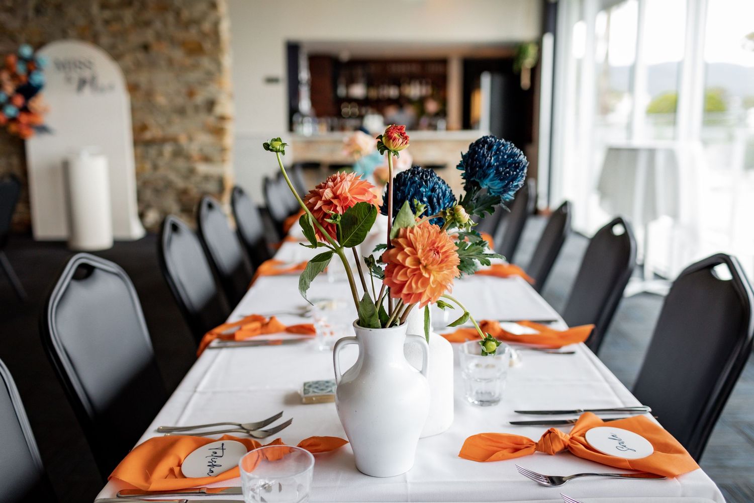 Long dining table set with orange napkins and floral centerpieces in white vases.