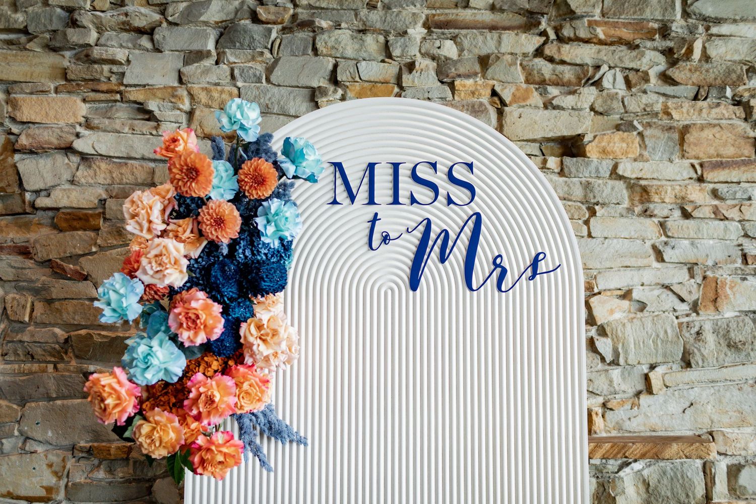 'Miss to Mrs' sign decorated with coral and blue florals against a stone wall.