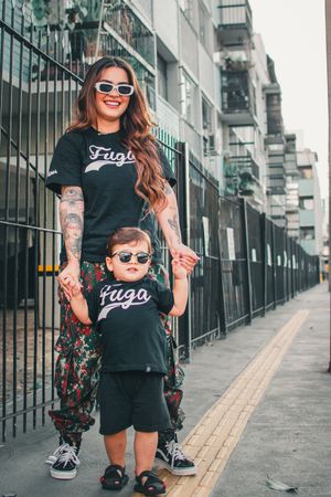 Mother and child in matching black t-shirts pose on urban sidewalk with coordinated streetwear looks and sunglasses.