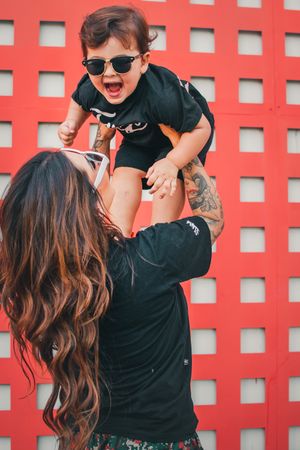 Joyful moment captured against red wall as parent lifts laughing child wearing sunglasses.