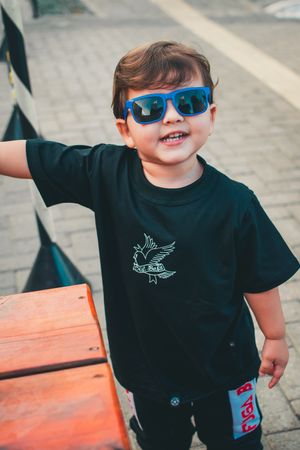 Happy child in black t-shirt and blue sunglasses smiles while standing next to orange bench.