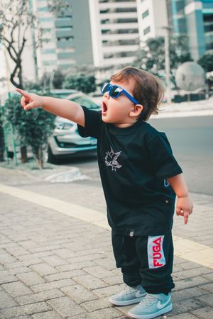 Toddler in streetwear and blue sunglasses points while standing on urban sidewalk.