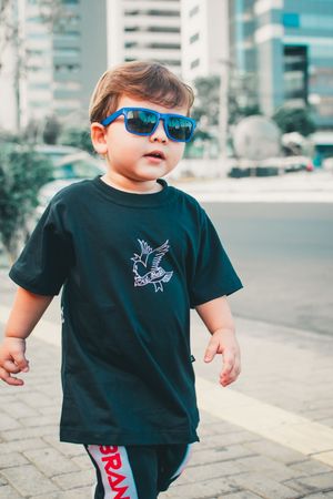 Child in black t-shirt and blue sunglasses poses confidently against urban backdrop.