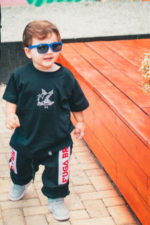 Smiling toddler in blue sunglasses and black outfit stands by orange bench with graffiti background.