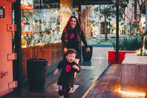 Parent and child in matching dark clothes with pink designs walking past urban storefronts.