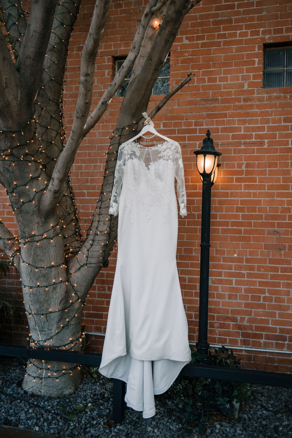 A white lace wedding dress hangs on display against a brick wall with string lights.
