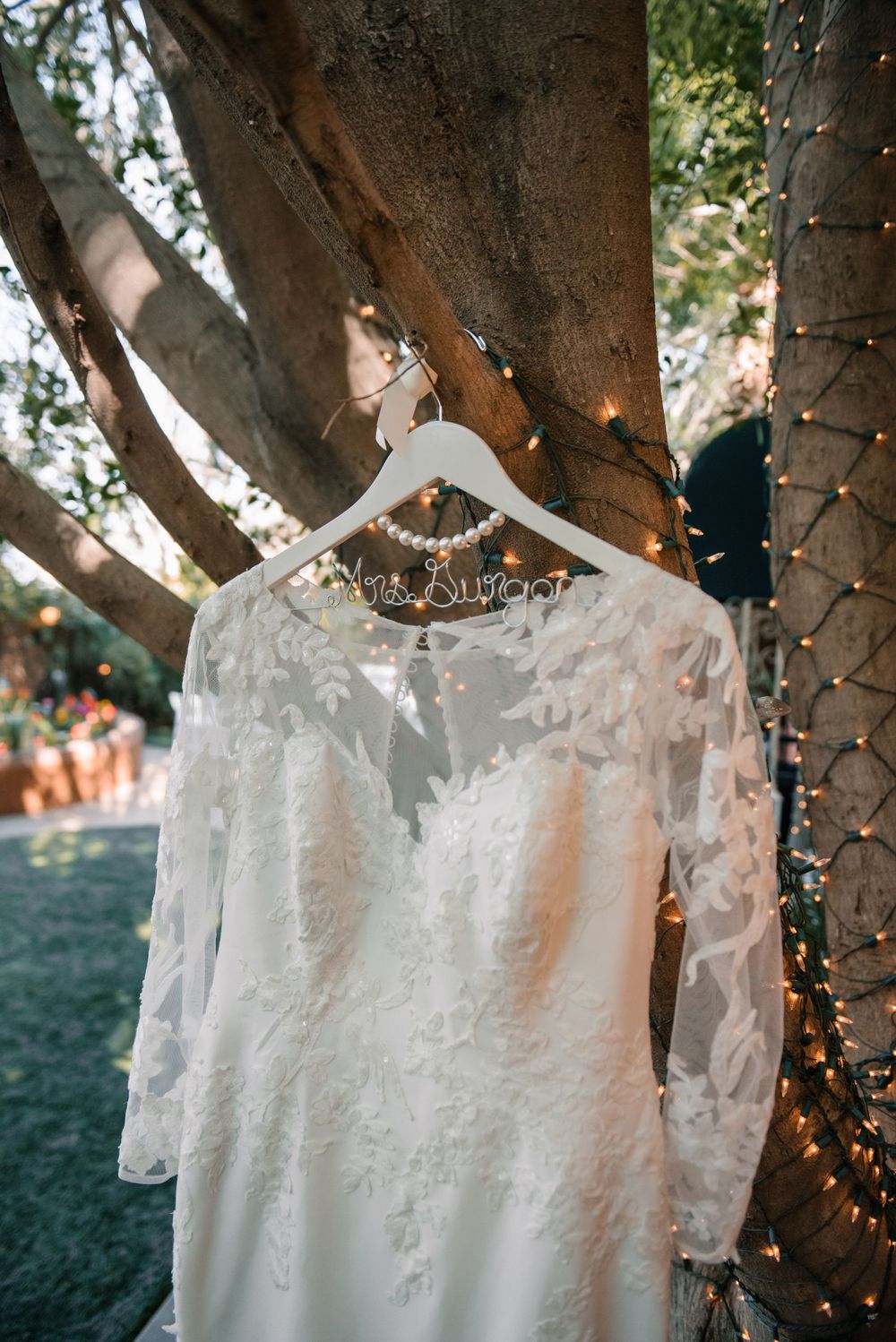 A white wedding dress with lace details hangs from a tree decorated with twinkle lights.