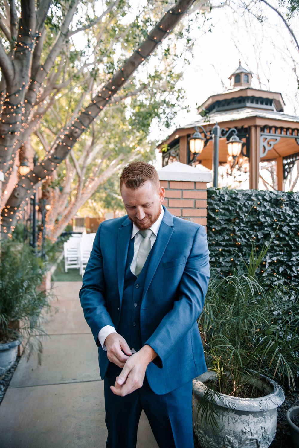 A person in a blue suit adjusts their cuffs while standing in an outdoor garden setting.