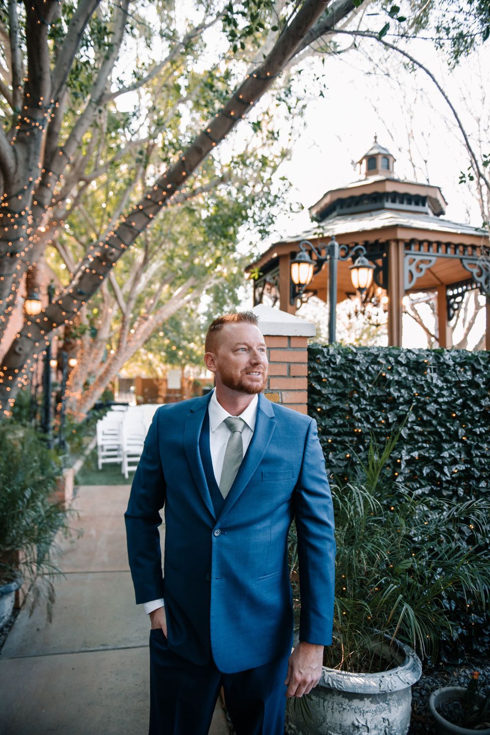 A person in formal attire stands in front of a brick wall and garden landscaping.