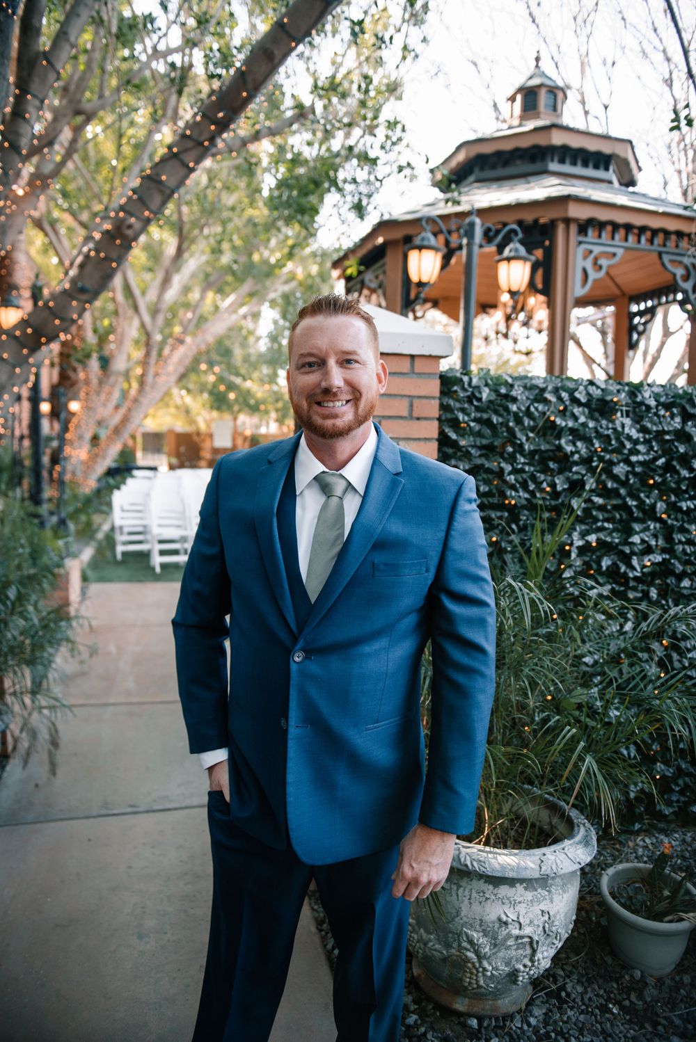 A person in a navy suit poses casually against a garden backdrop with brick walls and greenery.