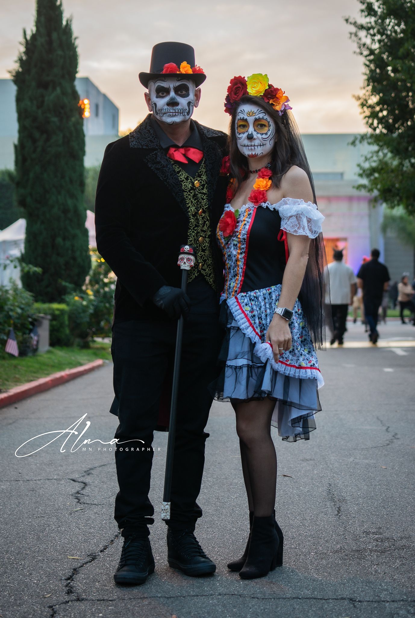 Couple in Day of the Dead costumes and skull makeup pose on evening street.