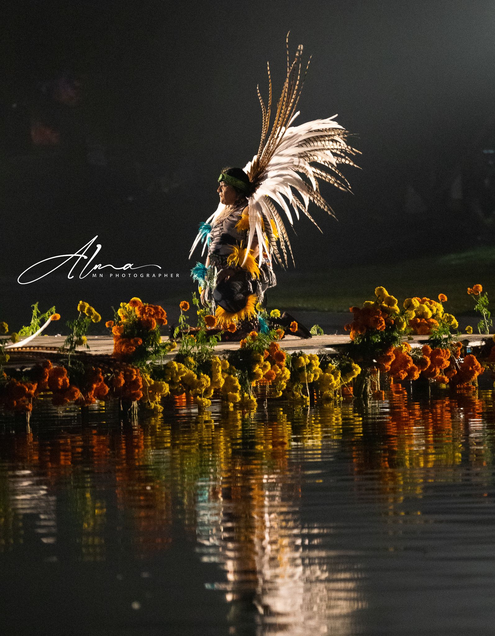 Performer in traditional Native American regalia with feather headdress dances over reflective water at night.
