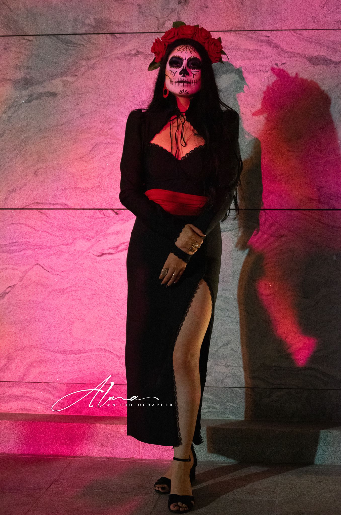 Figure in black dress with Day of the Dead makeup poses against pink-lit wall.